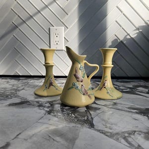May include: A set of three pale yellow ceramic pieces consisting of two matching candlestick holders and one small pitcher. Each item features a textured surface with hand-painted details of small brown birds perched on green branches with blue flowers.