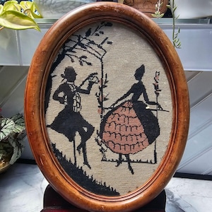 May include: An oval-shaped wooden frame containing a needlepoint embroidery piece. The artwork features black silhouettes of a person in a coat and a person in a wide skirt, set against a textured beige fabric background with black tree details.