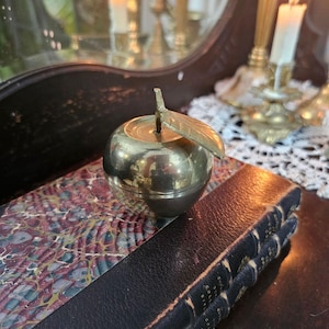 May include: A polished brass apple-shaped container with a removable lid and a single attached leaf. The metallic surface is reflective and smooth, featuring a rounded body and a stem that extends upward from the center of the top section.