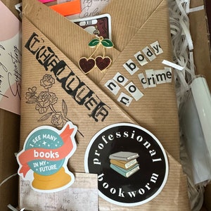 K&ouml;nnte beinhalten: Ein braunes Papierpaket mit verschiedenen Aufklebern und Verzierungen. Die Aufkleber beinhalten eine Kristallkugel mit dem Text "Ich sehe viele B&uuml;cher in meiner Zukunft", einen schwarzen Kreis mit dem Text "professioneller B&uuml;cherwurm" und einen Aufkleber mit dem Text "kein K&ouml;rper, kein Verbrechen".