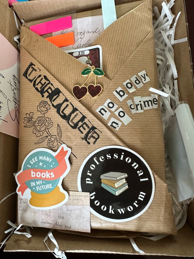 K&ouml;nnte beinhalten: Ein braunes Papierpaket mit verschiedenen Aufklebern und Verzierungen. Die Aufkleber beinhalten eine Kristallkugel mit dem Text "Ich sehe viele B&uuml;cher in meiner Zukunft", einen schwarzen Kreis mit dem Text "professioneller B&uuml;cherwurm" und einen Aufkleber mit dem Text "kein K&ouml;rper, kein Verbrechen".