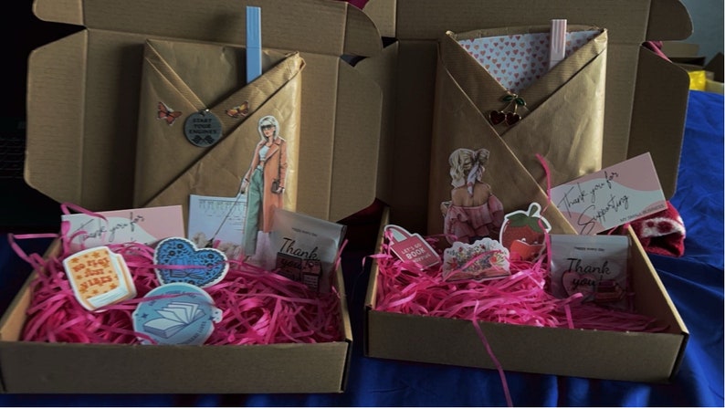 K&ouml;nnte beinhalten: Zwei ge&ouml;ffnete Kartons, gef&uuml;llt mit rosa zerknittertem Papier und verschiedenen kleinen Gegenst&auml;nden. Die Kartons enthalten Aufkleber, Karten und ein kleines Buch, das in braunes Papier eingewickelt ist. Die Kartons sind mit "Vielen Dank f&uuml;r Ihre Unterst&uuml;tzung" beschriftet.