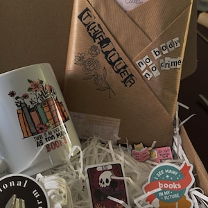 K&ouml;nnte beinhalten: Ein brauner Karton, gef&uuml;llt mit Geschenken zum Thema B&uuml;cher. Die Box enth&auml;lt eine wei&szlig;e Tasse mit einem Blumenmuster und dem Text "Es gibt nicht so etwas wie zu viele B&uuml;cher", eine Tarotkarte mit dem Text "Dunkle Romanze", einen Aufkleber mit dem Text "Ich sehe viele B&uuml;cher in meiner Zukunft" und einen Aufkleber mit dem Text "Kein K&ouml;rper, kein Verbrechen".