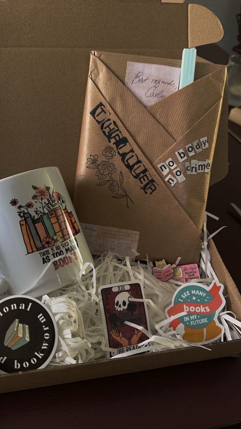 K&ouml;nnte beinhalten: Ein brauner Karton, gef&uuml;llt mit Geschenken zum Thema B&uuml;cher. Die Box enth&auml;lt eine wei&szlig;e Tasse mit einem Blumenmuster und dem Text "Es gibt nicht so etwas wie zu viele B&uuml;cher", eine Tarotkarte mit dem Text "Dunkle Romanze", einen Aufkleber mit dem Text "Ich sehe viele B&uuml;cher in meiner Zukunft" und einen Aufkleber mit dem Text "Kein K&ouml;rper, kein Verbrechen".