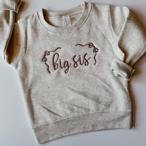 May include: A light grey sweatshirt featuring the embroidered words "big sis" in a cursive font, in dark brown thread. The sweatshirt has a crew neck, ribbed cuffs and hem. The sleeves are rolled up.