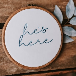 May include: A white embroidery hoop with a wooden frame, featuring blue embroidery that reads "he's here".