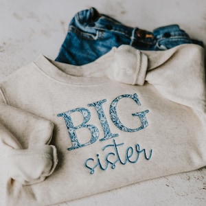May include: A cream-coloured long-sleeve sweatshirt featuring the text BIG in blue floral print and the word sister in blue cursive embroidery, paired with a pair of blue denim jeans folded behind the sweatshirt.
