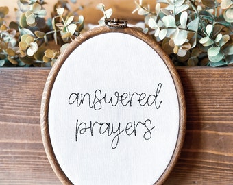 May include: A white embroidery hoop with a brown wooden frame. The hoop has the words "answered prayers" embroidered in black thread.