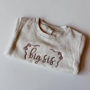 May include: A pale grey sweatshirt featuring the embroidered words "big sis" in a dark brown hue. The garment is neatly folded, highlighting the crew neck and the embroidered design.
