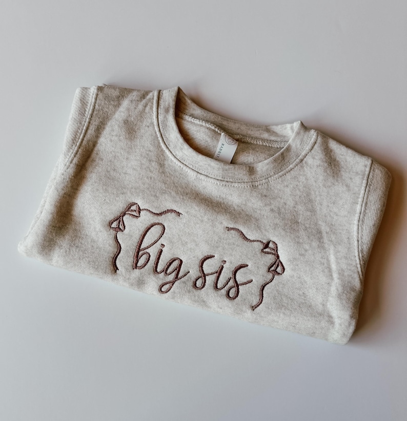 May include: A pale grey sweatshirt featuring the embroidered words "big sis" in a dark brown hue. The garment is neatly folded, highlighting the crew neck and the embroidered design.