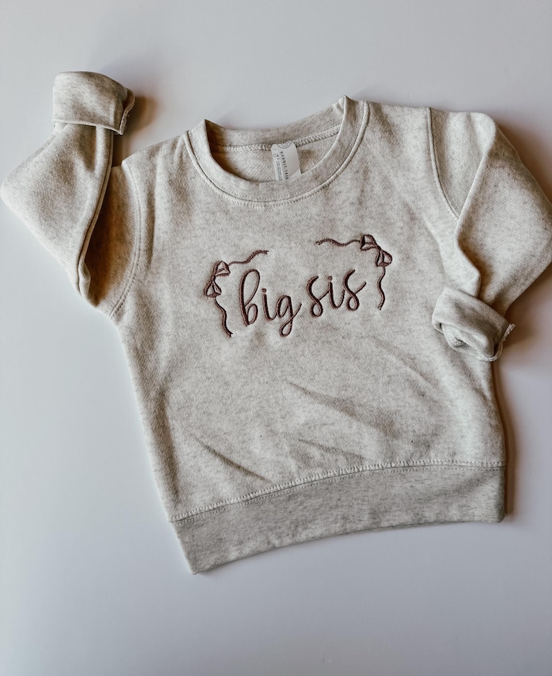 May include: A light grey sweatshirt featuring the embroidered words "big sis" in a cursive font, in dark brown thread. The sweatshirt has a crew neck, ribbed cuffs and hem. The sleeves are rolled up.