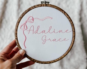 May include: Embroidered hoop art featuring the name "Adaline Grace" in pink script. A pink bow and ribbon design is stitched above the name. The embroidery is set in a wooden hoop with a decorative grain.