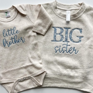 May include: A beige baby bodysuit with "little brother" embroidered in blue script next to a beige sweatshirt with "BIG sister" embroidered in blue floral letters and script. The clothing is made of a soft material.