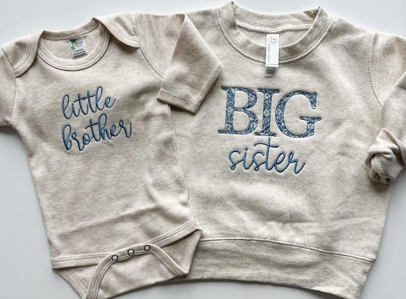 May include: A beige baby bodysuit with "little brother" embroidered in blue script next to a beige sweatshirt with "BIG sister" embroidered in blue floral letters and script. The clothing is made of a soft material.