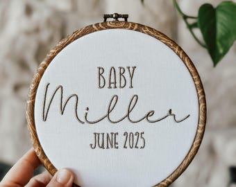 May include: A white embroidery hoop with brown wood trim. The hoop has the text "BABY Miller JUNE 2025" embroidered in brown thread.