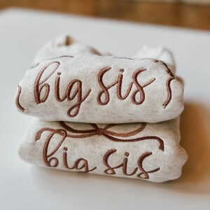 May include: Two folded, light grey sweatshirts stacked on top of each other. The top sweatshirt has the embroidered text "big sis" in a brown script. The bottom sweatshirt has the embroidered text "big sis" in a brown script.