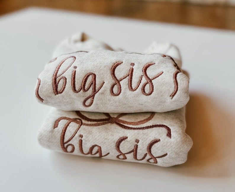 May include: Two folded, light grey sweatshirts stacked on top of each other. The top sweatshirt has the embroidered text "big sis" in a brown script. The bottom sweatshirt has the embroidered text "big sis" in a brown script.