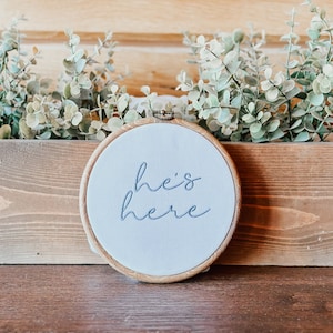 May include: A white embroidery hoop with a wooden frame, featuring the text "he's here" in blue embroidery thread.