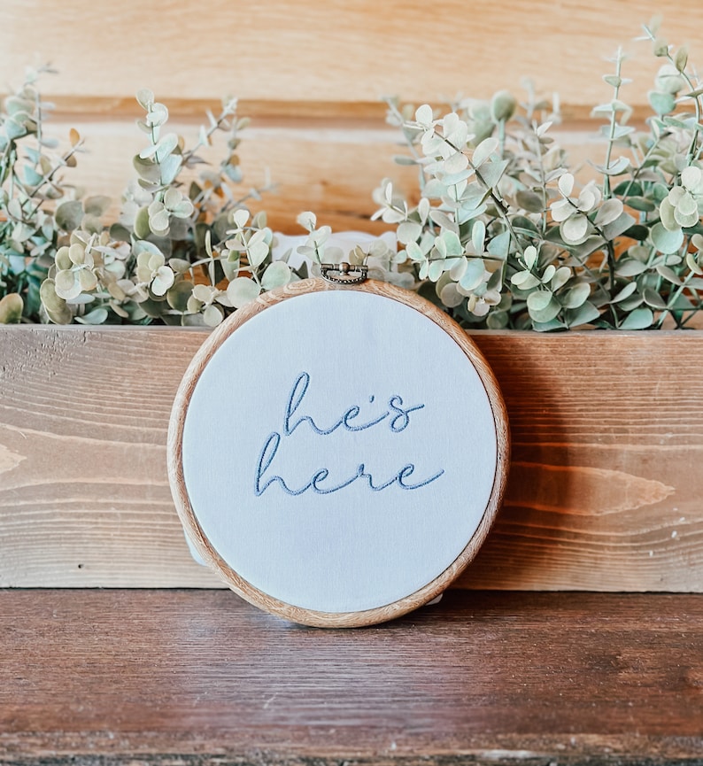 May include: A white embroidery hoop with a wooden frame, featuring the text "he's here" in blue embroidery thread.