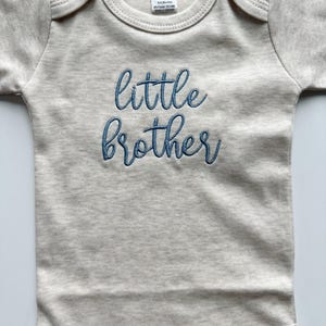 May include: A light beige baby bodysuit with short sleeves. The words "little brother" are embroidered in blue script across the front. The bodysuit has a round neckline and snap closures at the bottom.