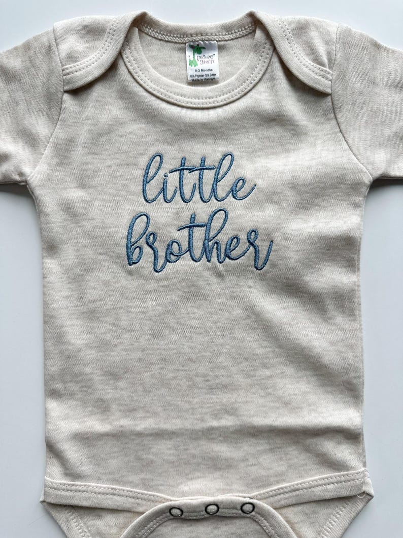 May include: A light beige baby bodysuit with short sleeves. The words "little brother" are embroidered in blue script across the front. The bodysuit has a round neckline and snap closures at the bottom.