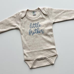 May include: A beige baby onesie with long sleeves. The words "little brother" are embroidered in blue script across the front. The onesie has a round neckline and snaps at the bottom.