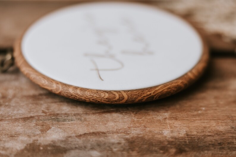 May include: Close-up of a round wooden embroidery hoop with a white fabric center. The fabric has the word "Thank you" written in cursive on it.