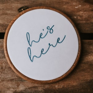 May include: A white embroidery hoop with a wooden frame. The hoop has blue embroidery that reads "he's here".