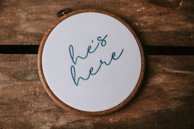 May include: A white embroidery hoop with a wooden frame. The hoop has blue embroidery that reads "he's here".