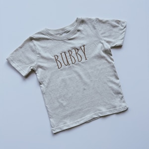 May include: A light beige short-sleeved t-shirt with the word "BUBBY" embroidered in brown thread.