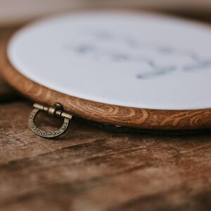 May include: A wooden embroidery hoop with a white fabric stretched across it. The hoop has a brass latch on the side.