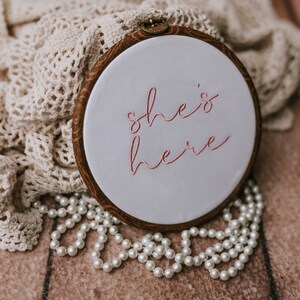 May include: A white embroidery hoop with a brown wooden frame. The hoop has pink embroidery that reads "she's here". The hoop is sitting on a brown surface with a white crocheted blanket and a string of white pearls.