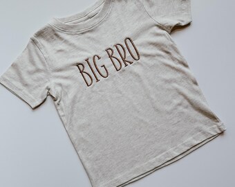 May include: A beige short-sleeved t-shirt with the words "BIG BRO" embroidered in brown on the front.