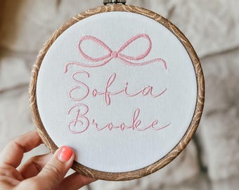 May include: Embroidered hoop with a pink bow and the names "Sofia Brooke" in pink cursive lettering.