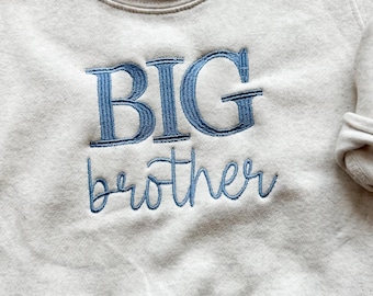 May include: A beige sweatshirt with the words "BIG brother" embroidered in blue thread. The sweatshirt has a crew neck and long sleeves. The text is in a bold, block font for "BIG" and a cursive font for "brother".