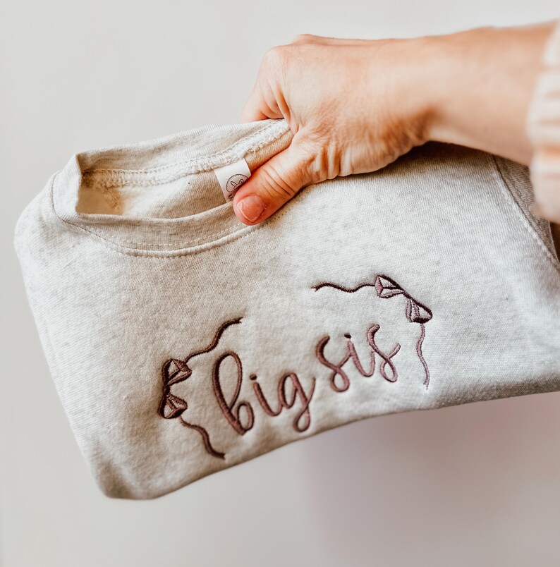 May include: A light grey sweatshirt featuring the embroidered words "big sis" in a cursive style. The design includes two kite-like shapes with string details. The sweatshirt is held up by a hand.
