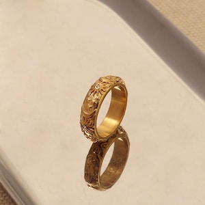 May include: A gold-colored ring with an engraved floral pattern and small, clear gemstones. The ring is reflected on a shiny surface, showcasing its intricate design. The ring's band features detailed carvings of flowers and other decorative elements.