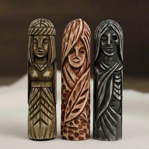 May include: Three carved wooden statues of female figures. The statues are in shades of brown and grey, with detailed carvings of faces, hair, and clothing. The central figure has a leaf design, while the others have geometric patterns. The statues are displayed on a white surface.