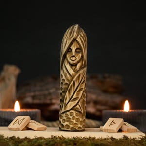 May include: A carved wooden statue of a woman with closed eyes and flowing hair. The statue is light brown and features intricate details, including leaf and dot patterns. Wooden runes and lit candles are in the background.