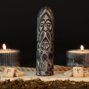 May include: A carved wooden statue of a bearded figure with runes, in shades of gray. The statue is the focal point, with lit candles on either side. Wooden runes and moss are in the foreground, with a rope in the background.
