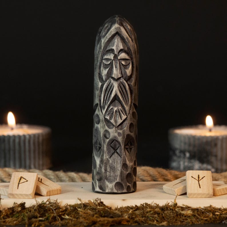 May include: A carved wooden statue of a bearded figure with runes, in shades of gray. The statue is the focal point, with lit candles on either side. Wooden runes and moss are in the foreground, with a rope in the background.