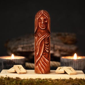 Op de afbeelding: Een roodbruin houten beeld van een figuur met een skeletachtig gezicht en lang haar. Het beeld staat op een houten voet met runentegels. Brandende kaarsen op de achtergrond creëren een mystieke sfeer. Het beeld is gedetailleerd gesneden.
