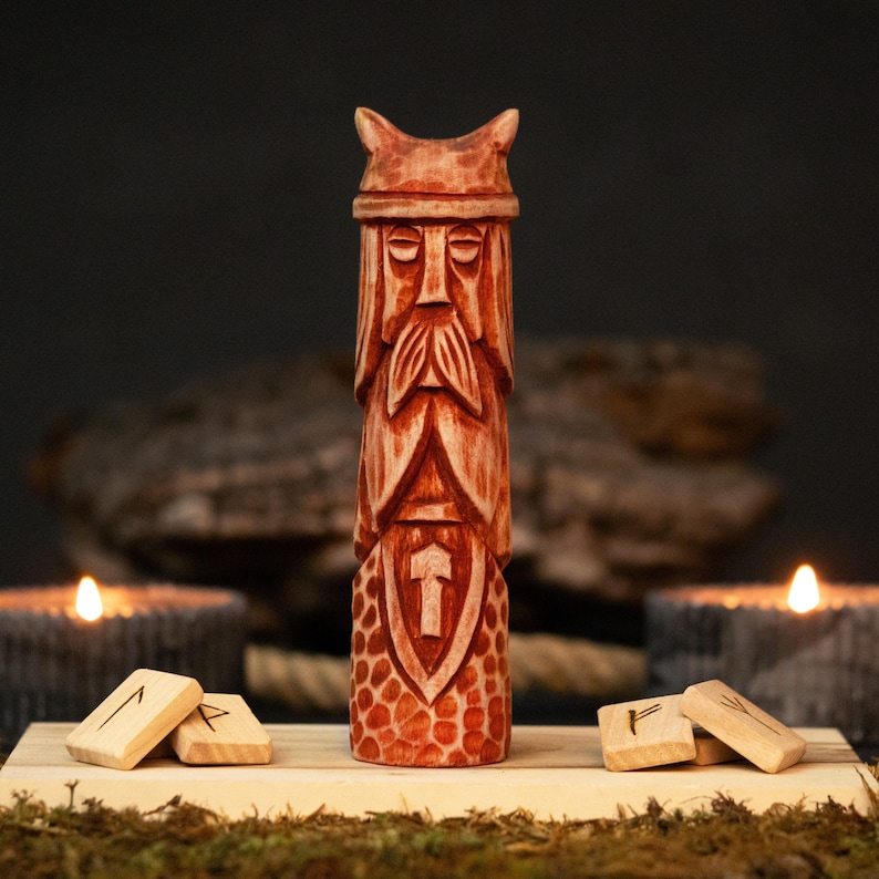 May include: A carved wooden statue of a bearded figure with a horned helmet. The statue is reddish-brown and features intricate details. A shield with a hammer is carved on the chest. Wooden runes and lit candles are in the background. The statue is approximately 10 inches tall.