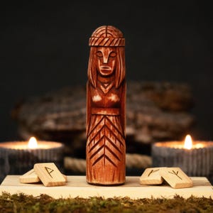 Puede incluir: Una estatua de madera tallada de una mujer con un tocado detallado y cabello suelto. La estatua es de color marrón cálido y se encuentra sobre una base de madera con varias pequeñas piedras rúnicas de madera. Dos velas encendidas están en el fondo.