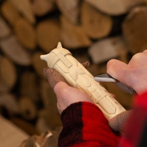 May include: A person carving a light-colored wooden object with a knife. The object has a detailed, geometric design. The person is wearing a red and black plaid shirt and a brown fingerless glove.