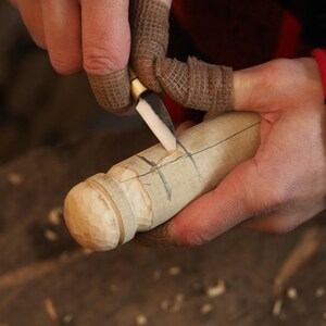 May include: A close-up of a person carving a wooden object with a small knife. The object is cylindrical with a rounded, textured end. The person is wearing finger protectors. The wood has pencil markings.