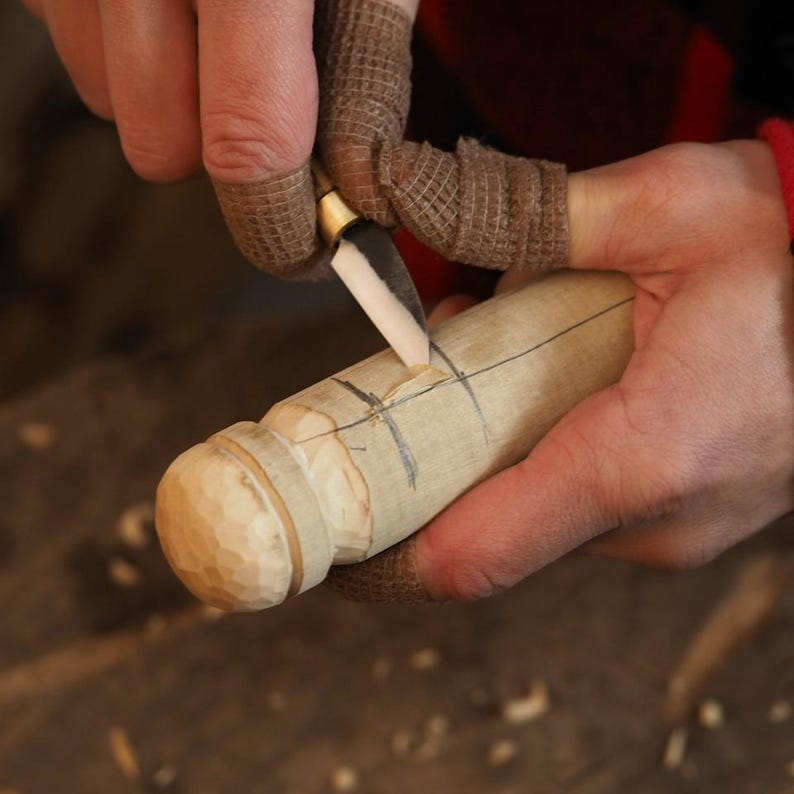 May include: A close-up of a person carving a wooden object with a small knife. The object is cylindrical with a rounded, textured end. The person is wearing finger protectors. The wood has pencil markings.