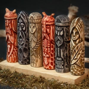 May include: A collection of six carved wooden figures, each depicting a stylized face with intricate details. The figures vary in color, ranging from reddish-orange to dark gray and natural wood tones. They are arranged on a wooden base, showcasing a variety of carved patterns and textures.