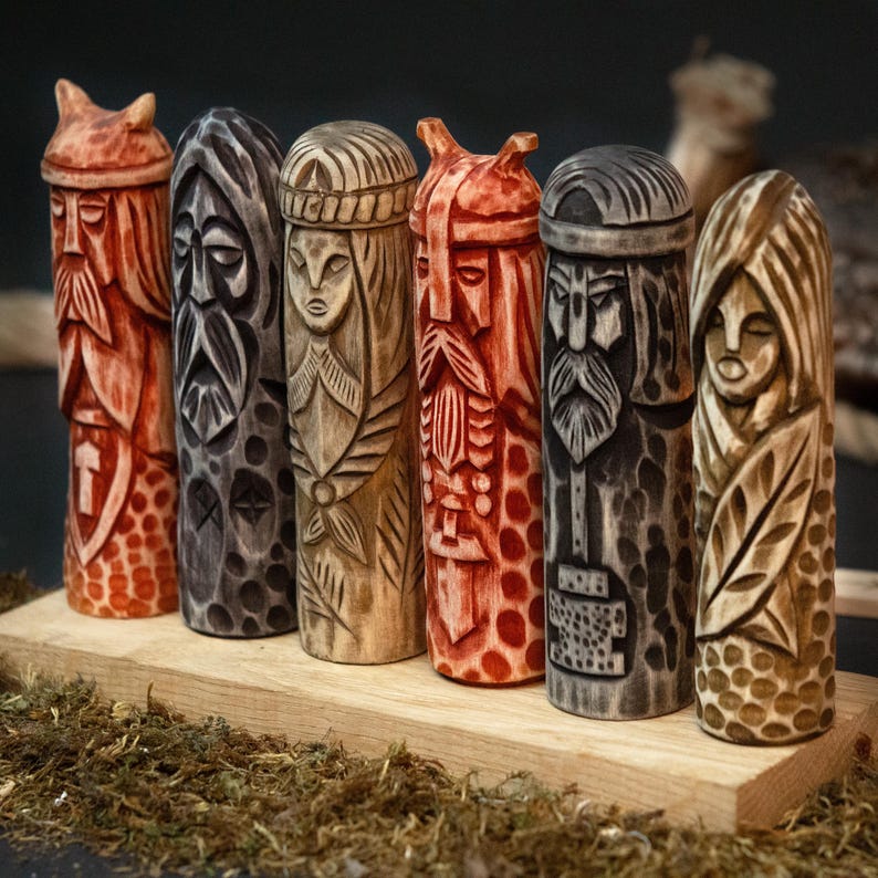 May include: A collection of six carved wooden figures, each depicting a stylized face with intricate details. The figures vary in color, ranging from reddish-orange to dark gray and natural wood tones. They are arranged on a wooden base, showcasing a variety of carved patterns and textures.
