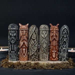 May include: Six carved wooden figures with detailed faces and intricate designs. The figures are in shades of gray, brown, and beige, and are arranged on a wooden base with moss. Two lit candles are in the background.